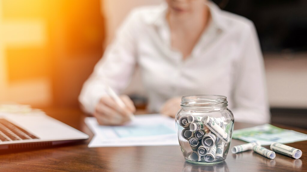 Jar with rolled banknotes table laptop papers woman background